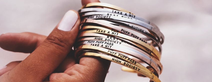 Puff Puff Pass Bracelets Eine Hand hält einen Stapel goldener und silberner Armbänder mit unterschiedlichen eingravierten Sätzen.