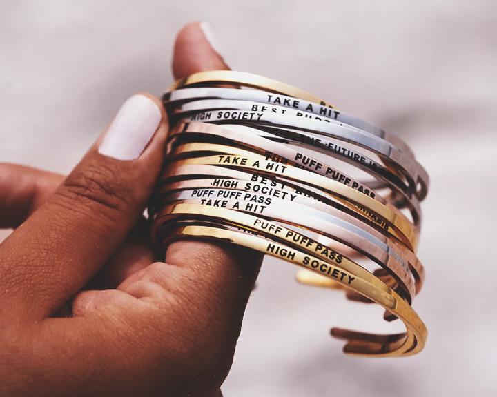 Puff Puff Pass Bracelets Eine Hand hält einen Stapel goldener und silberner Armbänder mit unterschiedlichen eingravierten Sätzen.