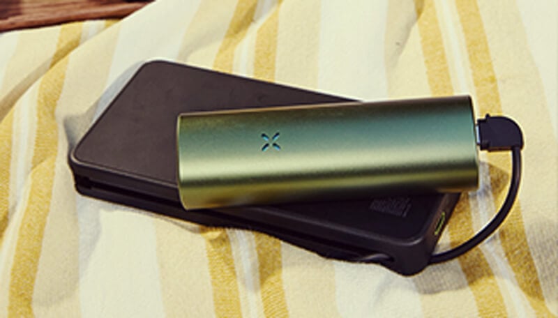 Aufladen des Pax Flow Vaporizers Ein schlankes gRüneS pax flow vaporizer-gerät, das über ein usb-kabel mit einem schwarzen powERBank-ladegerät verbunden ist und auf einem gelb-weißen gestreiften handtuch für unterwegs bequemes aufladen ruht.