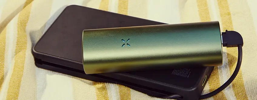 Aufladen des Pax Flow Vaporizers Ein schlankes grünes Pax Flow Vaporizer-Gerät, das über ein USB-Kabel mit einem schwarzen Powerbank-Ladegerät verbunden ist und auf einem gelb-weißen gestreiften Handtuch für unterwegs bequemes Aufladen ruht.