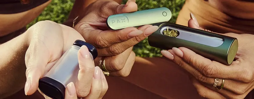 Befüllen des Pax Flow Vaporizers Zwei Hände, die zusammen den Pax Flow Vaporizer mit gemahlenem Cannabis aus einem kleinen Grinder füllen, und den einfachen Ladeprozess im Freien auf grünem Gras demonstrieren.