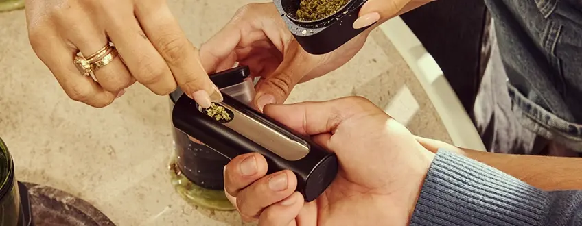 Nachfüllen des Pax Flow Vaporizers Eine Hand, die die Kammer des Pax Flow Vaporizers mit frischem gemahlenem Cannabis aus einer kleinen Schaufel eines Grinders nachfüllt, auf einem Marmortisch für präzises Nachladen platziert.