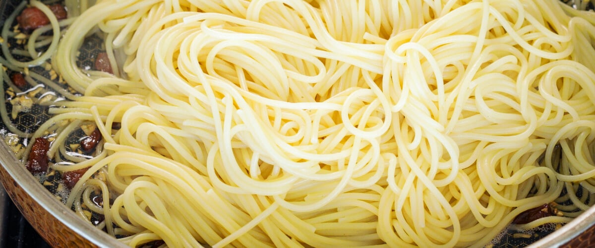 Abgetropfte Spaghetti werden mit knuspriger Pancetta und infundierter Butter in der Pfanne vermengt, vierter Schritt für eine Cannabis-Carbonara